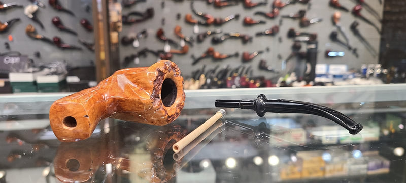 Brigham - Designer Series - A smoking pipe with a wooden body and a black mouthpiece, displayed on a glass shelf with a collection of pipes in the background.