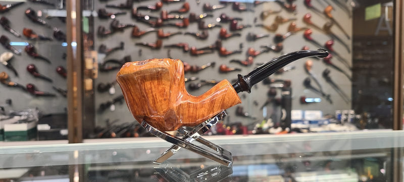 Brigham - Designer Series - A smoking pipe with a wooden body and a black mouthpiece, displayed on a glass shelf with a collection of pipes in the background.

Details