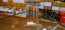 A clear acrylic display stand holding several brown cylindrical packaged items, likely cigars or cigar-related products, with visible labels. The stand is placed on a wooden counter inside a retail or warehouse setting, with shelves, boxes, and other products blurred in the background.