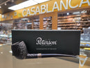A dark brown textured smoking pipe with a dark handle and a silver band, displayed in front of its black Peterson of Dublin box on a reflective surface, with a blurred shop interior in the background.