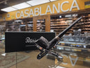 A black smoking pipe with a textured bowl and polished silver metal stem, displayed on a stand in front of its black branded box. The background shows a blurred shop interior with a prominent yellow sign reading 'CASABLANCA'.