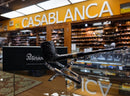 A black smoking pipe with a textured bowl and gold-toned band, displayed on a clear stand in front of a black Peterson box inside a retail shop. A blurred background shows shelves and a large yellow 'CASABLANCA' sign.