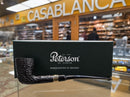 A black Peterson of Dublin smoking pipe with a textured briar stem and gold band, displayed in front of its dark green branded box on a glass display counter inside a shop
