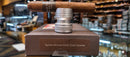 A single brown cigar resting horizontally on a silver metal ring, displayed on top of a brown box labeled 'Ramon Allones Small Club Coronas' in a blurred cigar store setting.