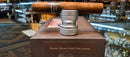 A single Ramon Allones cigar resting horizontally on a metallic silver display stand atop a brown presentation box labeled 'Ramon Allones Small Club Coronas,' with a blurred cigar shop interior in the background.