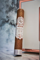 A single brown cigar with a white and rose gold label reading 'RP White Label' stands upright on a marble surface with smoke in the background and a framed mirror behind it.