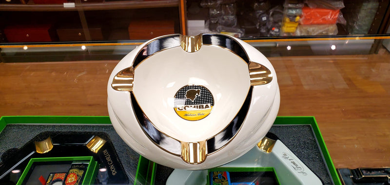 A Cohiba Premium Ashtray displayed on a glass shelf in a store, with a gold and black color scheme and the Cohiba logo visible.
