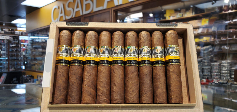 A wooden humidor box containing Cohiba Maduro 5 - Magicos cigars, with visible cigar labels and branding.