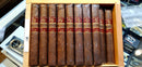 A wooden humidor box containing multiple Rocky Patel cigars, with individual cigars visible in slots.

